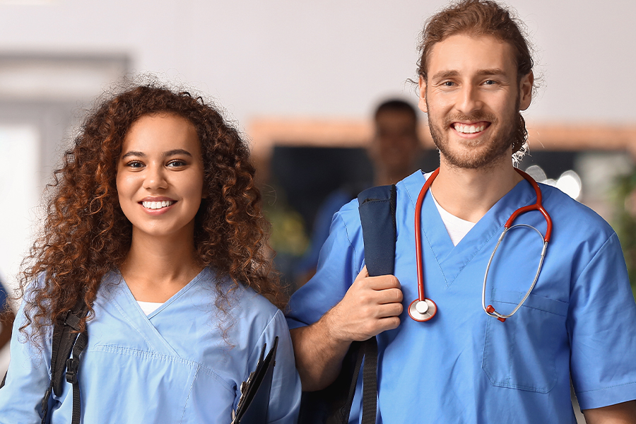 Two nursing students in blue scrubs smile and carry backpacks in a healthcare facility hallway. One wears a stethoscope around their neck.