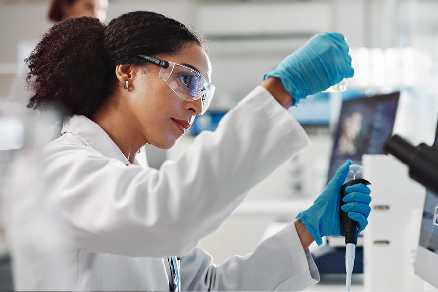 A scientist in a white lab coat, safety glasses, and blue gloves uses a pipette to transfer a sample in a laboratory setting.