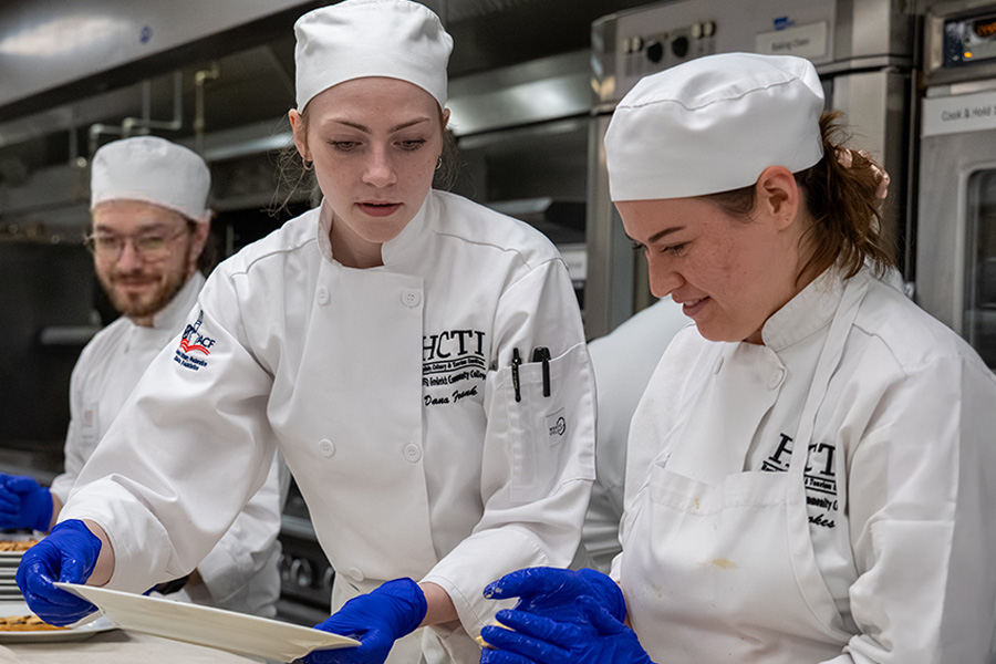 Three culinary students in chef coats plate food together in a commercial kitchen