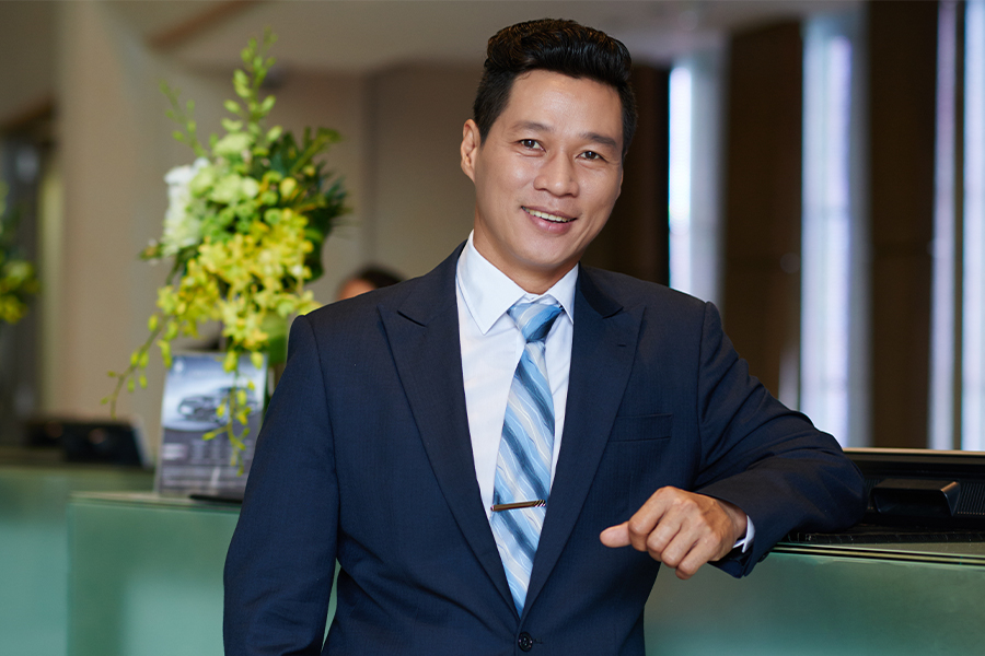 A smiling hospitality professional in a navy suit and striped tie leans against a front desk counter in a hotel lobby, with a floral arrangement visible in the background.